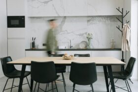 Przy stole jadalnianym dzieje się zdecydowanie więcej niż tylko jedzenie posiłków. To miejsce, gdzie odrabia się lekcje, pije poranną kawę, rozkłada dokumenty z pracy, pakuje świąteczne prezenty, a czasem gra w planszówki do późnej nocy. Nic dziwnego, że stół szybko przestaje być wyłącznie meblem do jedzenia, a staje się centrum domowego życia – tym bardziej w mieszkaniach, gdzie jadalnia jest częścią otwartej strefy dziennej. Na zdjęciu w katalogu każdy stół wygląda dobrze. Problemy zaczynają się dopiero wtedy, gdy okazuje się, że po dosunięciu krzeseł nie da się swobodnie przejść, przy krótszym boku nikt nie chce siedzieć, bo nogi stołu przeszkadzają, a rozkładanie blatu wymaga przesuwania pół pokoju. Z drugiej strony zbyt mały stół wygląda nieproporcjonalnie w przestrzeni, nie wykorzystuje potencjału wnętrza i sprawia, że większe spotkania kończą się dokładaniem tymczasowych stolików. Dobór stołu do jadalni to więc nie tylko kwestia gustu, ale przede wszystkim proporcji, ergonomii i dopasowania do tego, jak naprawdę korzystasz z przestrzeni. Liczy się liczba domowników, metraż, układ pomieszczenia, a nawet to, jak często zapraszasz gości. W tym artykule przeprowadzimy Cię przez najważniejsze decyzje: od określenia potrzeb, przez dobór wymiarów i kształtu, aż po materiały i styl, które sprawią, że stół będzie wygodny na co dzień i spójny z resztą wnętrza.